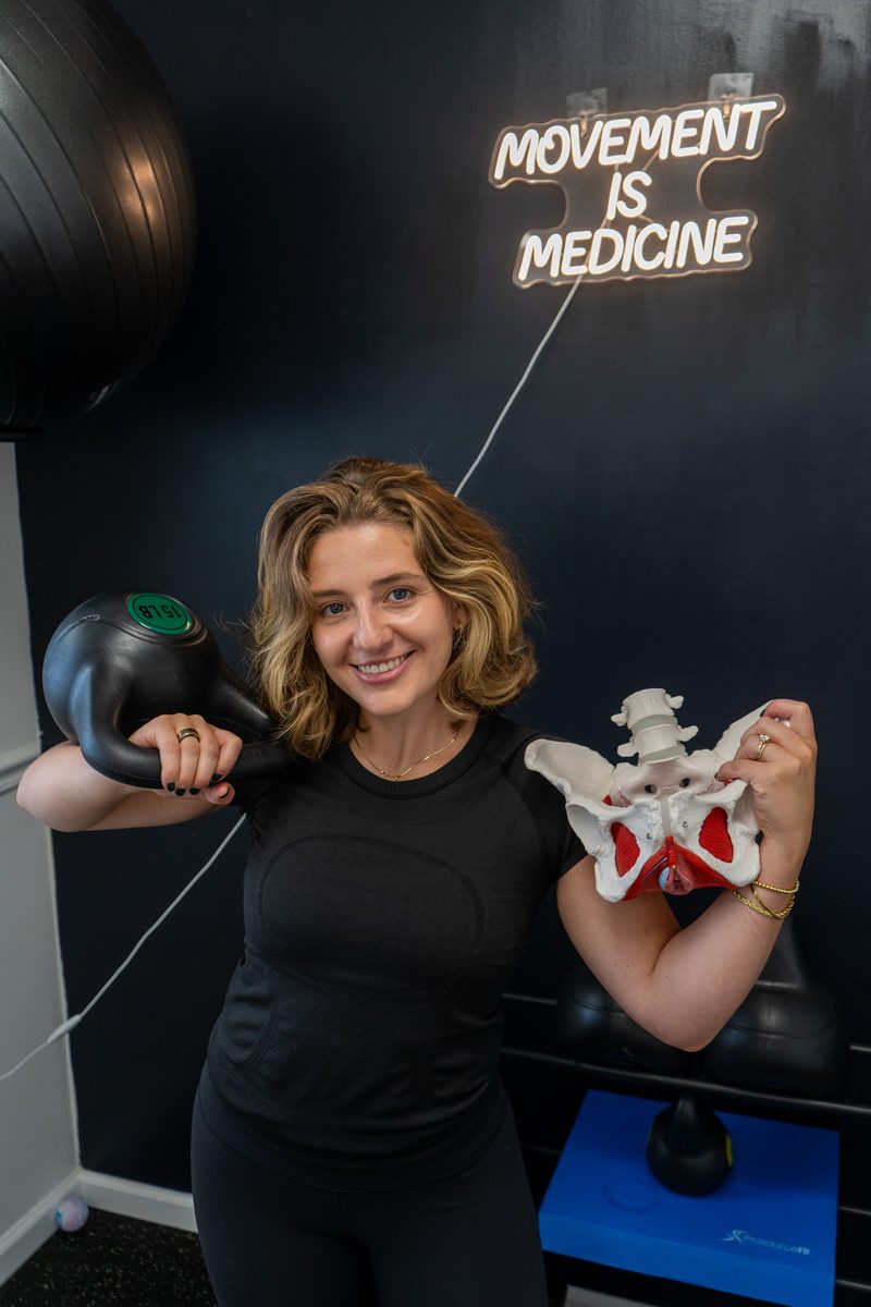 Erica Bermudez, OTR/L, holding a kettlebell and a pelvic model under a Movement Is Medicine sign