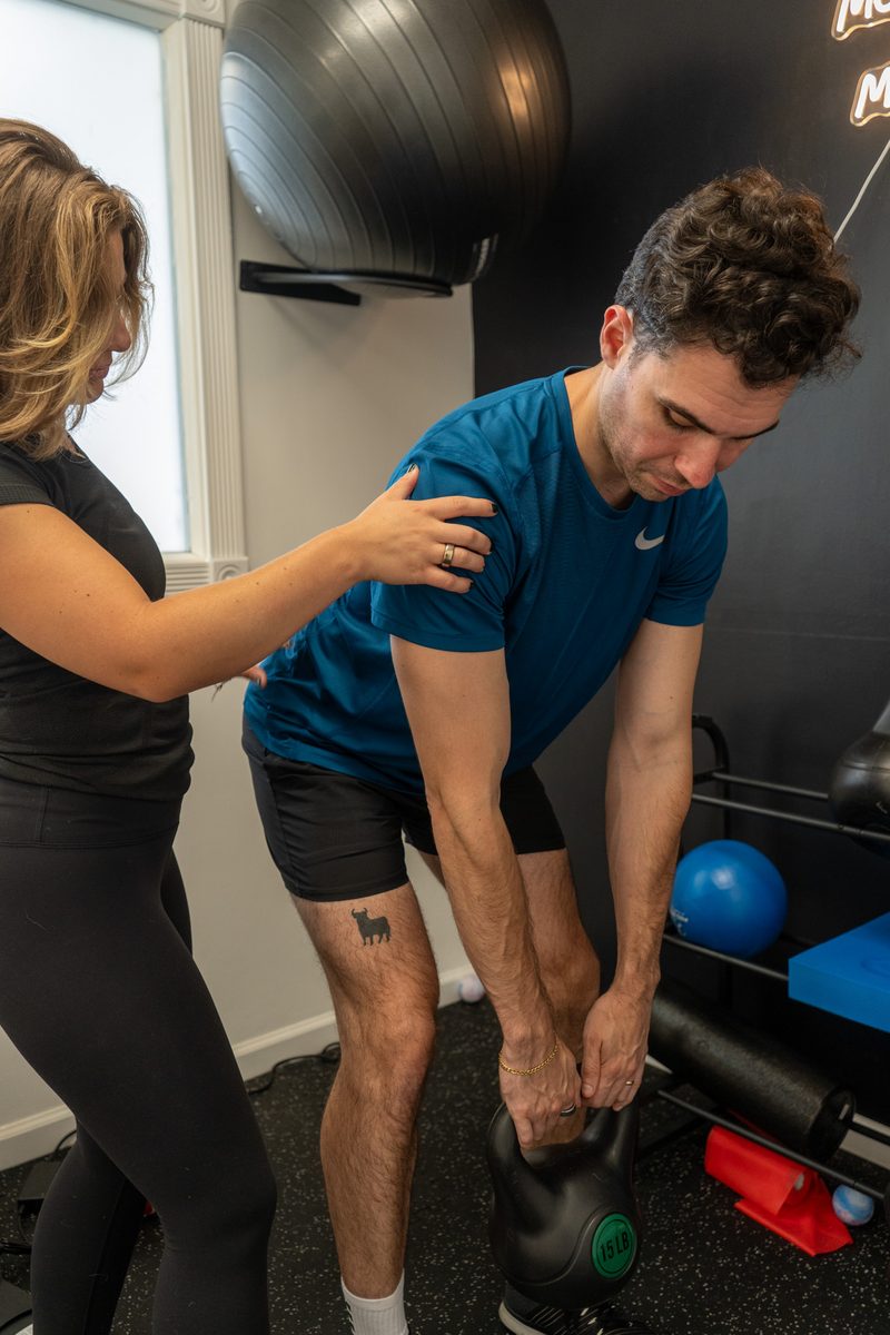Erica coaching a male client through a kettlebell deadlift, cueing his back position